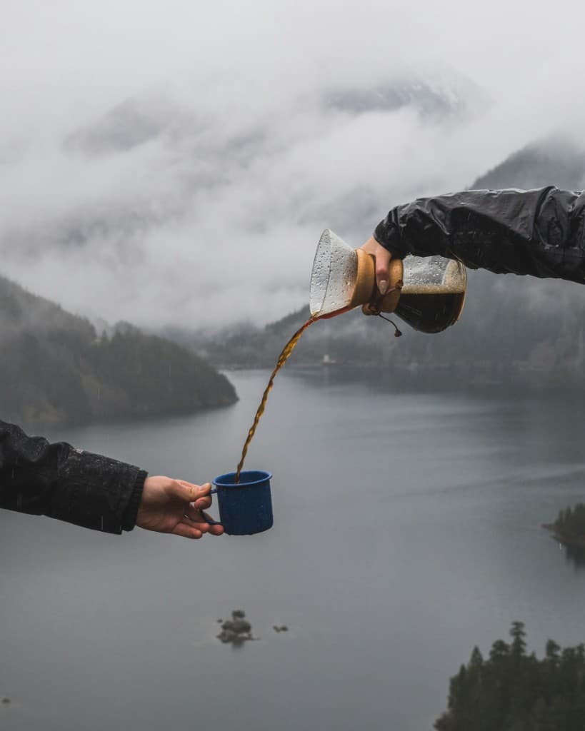 Pouring coffee from a Chemex into a blue enamel camp mug outdoors, misty mountains and water beyond