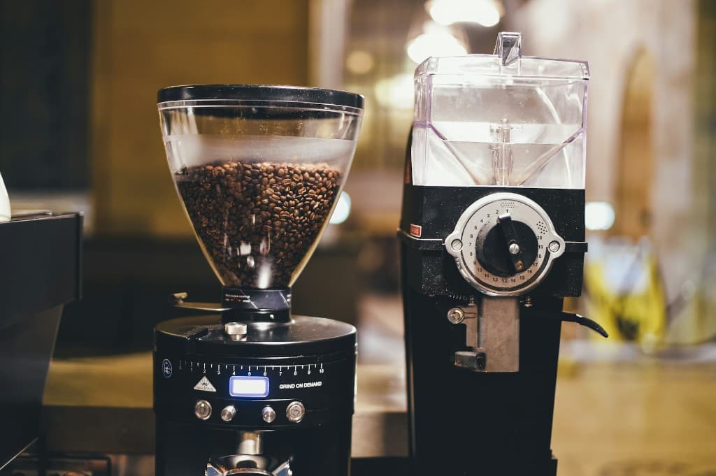 Two professional burr coffee grinders on a cafe counter — an espresso grind-on-demand grinder with beans in the hopper and a larger shop grinder with an adjustment dial