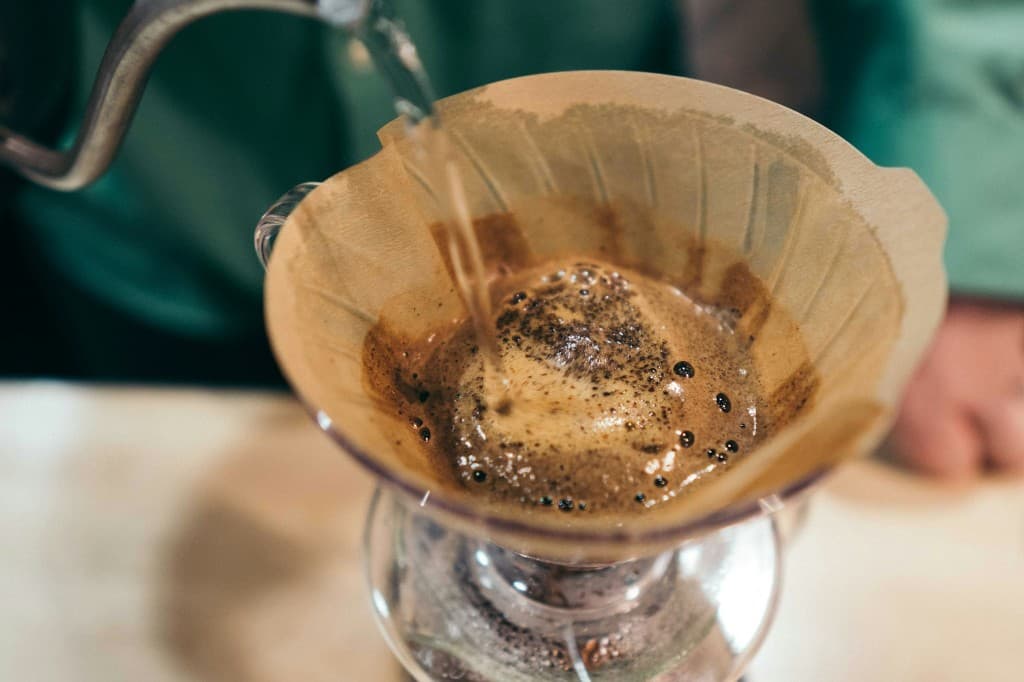 Gooseneck kettle pouring a stream of water into a V60 cone, coffee grounds blooming with foam beneath the pour